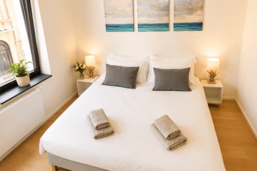 Chambre moderne à Mattbnb à Luxembourg avec lit queen-size, oreillers gris, serviettes pliées, œuvres marines et éclairage chaleureux