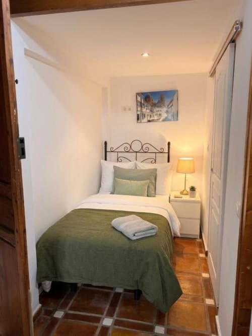 Acogedora habitación doble en Hotelito Oliva con cama de hierro forjado, colcha verde, lámpara y arte con paisaje urbano europeo