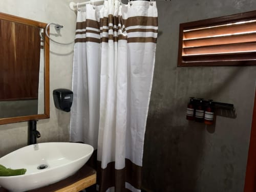 Baño moderno del hostal Cayuco Maya en Bacalar con lavamanos blanco, cortina de ducha a rayas marrones y detalles de madera