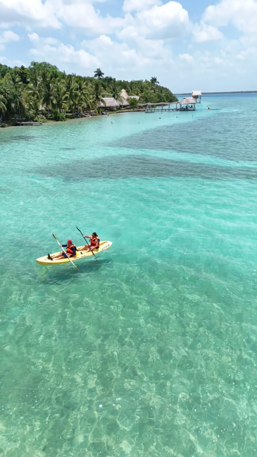 Dos huéspedes navegando en kayak en la laguna turquesa de Bacalar con resort de techos de palapa y palmeras de fondo