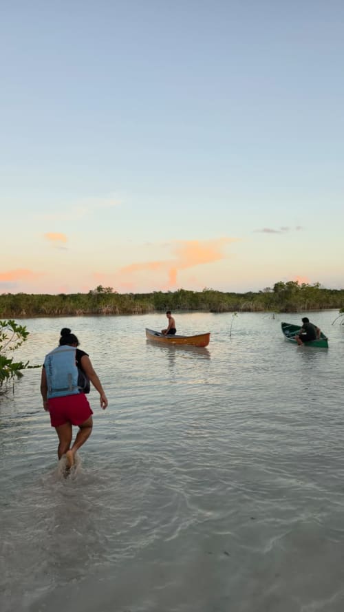 Persona adentrándose en las tranquilas aguas de la laguna al atardecer con botes de madera y manglares, Bacalar México