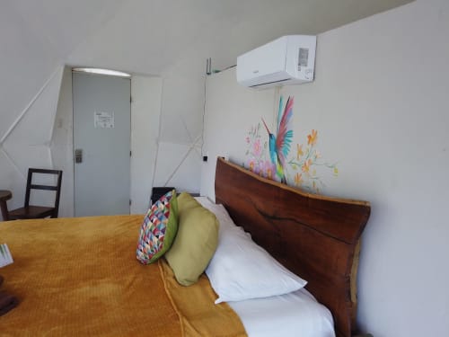 Acogedora habitación en el hostel Cayuco Maya de Bacalar con cama de madera, almohadones coloridos, aire acondicionado y arte floral en la pared.