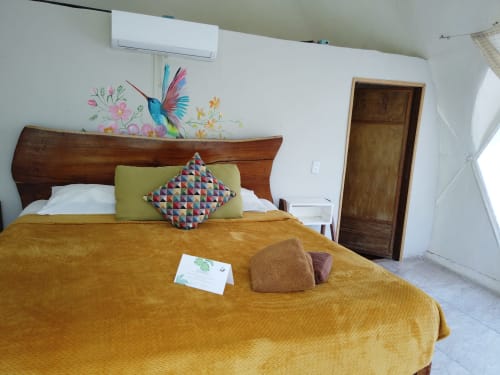 Dormitorio colorido en hostel Cayuco Maya en Bacalar con arte mural de colibrí, cama de madera y ropa de cama amarillo mostaza