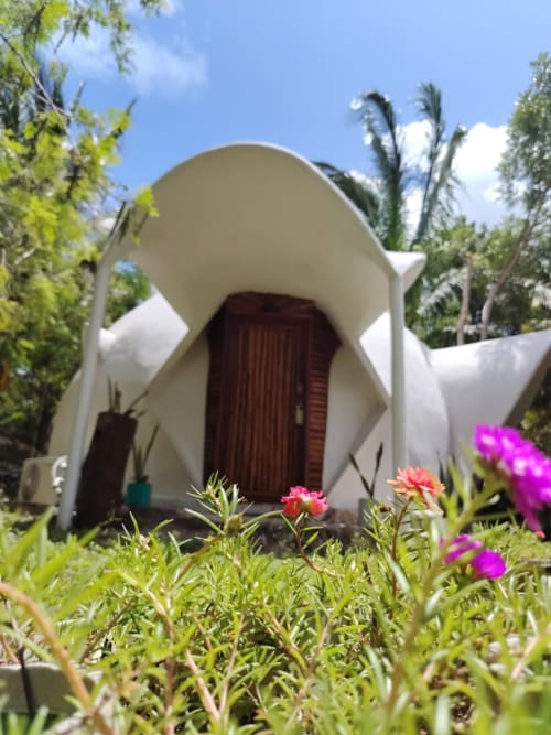 Domo geodésico blanco con puerta roja rodeado de flores rosas y plantas tropicales en el hostal Cayuco Maya de Bacalar, México