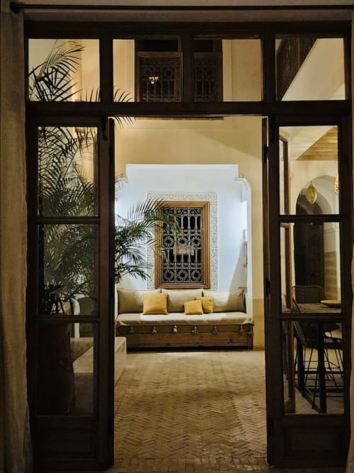 Portes en bois sombre s'ouvrant sur l'intérieur élégant du Riad Casa Angelo avec plantes en palmier, coussins jaunes sur banquette et fenêtre ornementale