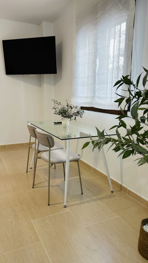 Modern studio apartment workspace in Baeza with glass table, upholstered chair, wall-mounted TV, and natural light from