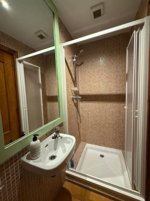 Baño moderno en Hotelito Oliva Pensión con paredes de azulejos marrones, lavabo blanco, espejo y ducha con puertas blancas.