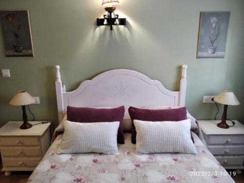 Dormitorio acogedor en estudio Casa Lahuerta con cabecero blanco, almohadas burdeos, ropa de cama floral, lámparas gemelas y pale