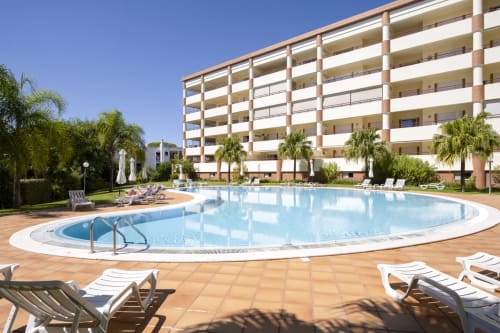 RB Algarve hotel in Vilamoura with curved swimming pool, white loungers, palm trees, and modern apartment building under