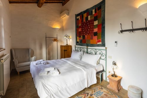 Camera da letto rustica con biancheria bianca, letto in ferro verde, coperta variopinta, travi di legno e muri in pietra a Buonconvento