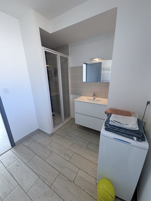 Salle de bain moderne avec vasque blanche, miroir, douche et lave-linge à la Résidence des dunes à Landéda