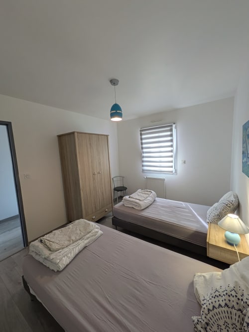 Chambre lumineuse à la Résidence des dunes avec deux lits simples, armoire en bois, stores à rayures et luminaire bleu suspendu