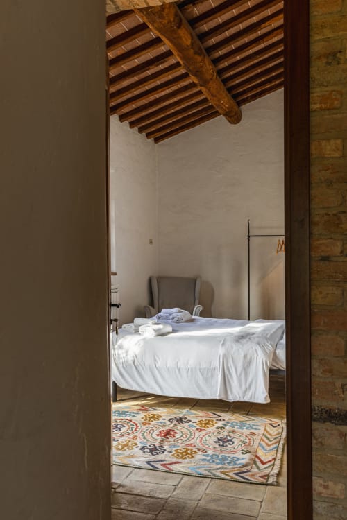 Camera da letto con biancheria bianca, travi in legno a vista e tappeto decorato all'hotel Quarantallina di Buonconvento, Italia