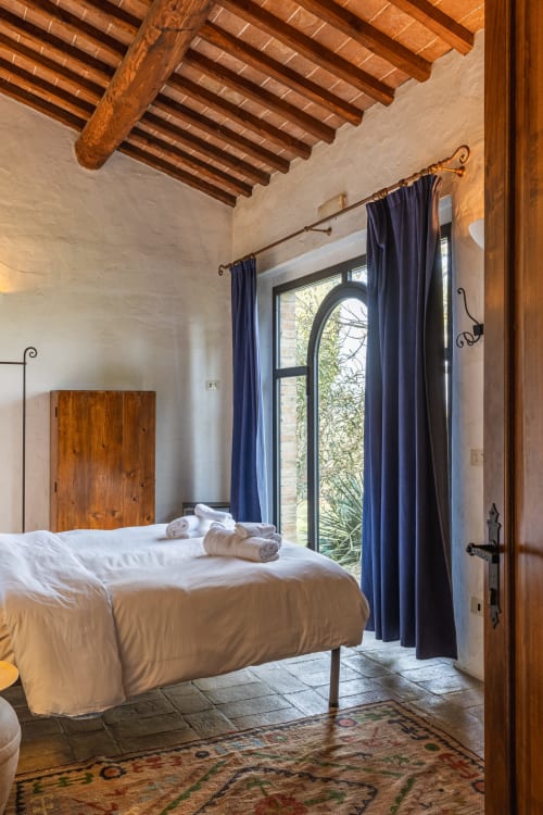 Camera da letto rustica a Buonconvento con travi in legno a vista, pareti bianche, tende blu e porte con vetri ad arco