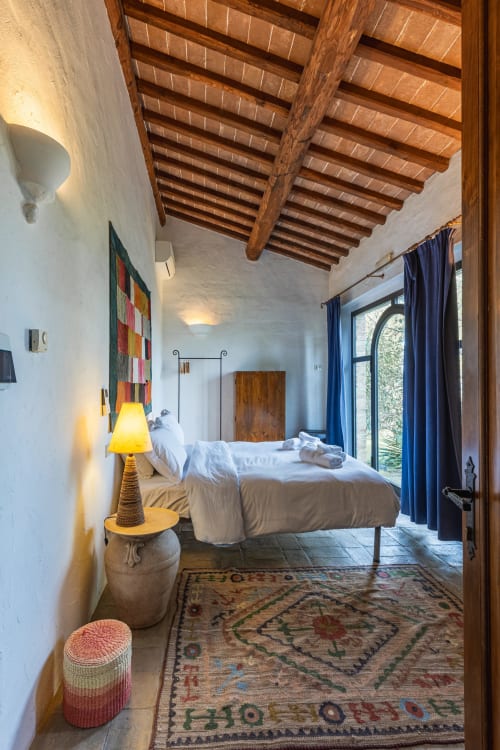 Camera rustica toscana con travi in legno a vista, pareti bianche, tende blu e tappeto tradizionale a Buonconvento
