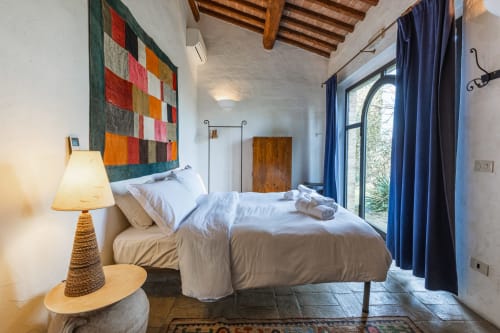 Camera luminosa dell'hotel Buonconvento con letto queen, trapunta colorata, travi in legno, tende blu e pavimento in pietra