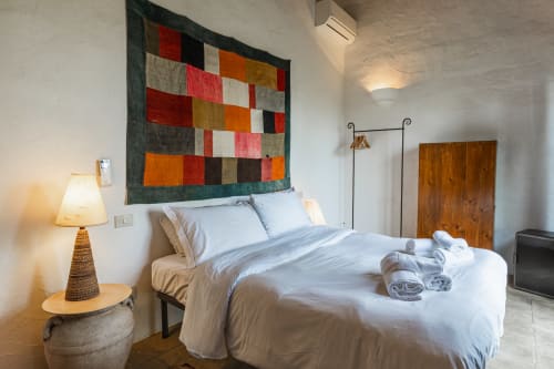 Camera da letto con opere d'arte patchwork colorate, biancheria bianca, lampada e mobili in legno nell'appartamento hotel di Buonconvento