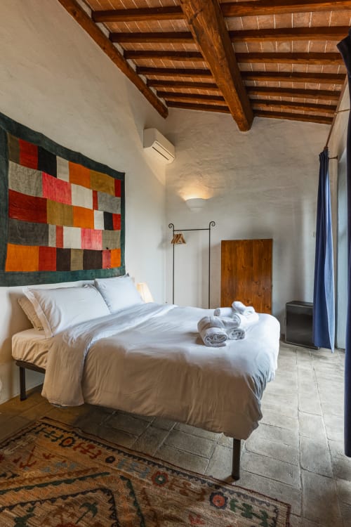 Camera da letto rustica con travi in legno, trapunta colorata, biancheria bianca e pavimento in cotto presso l'hotel Buonconvento