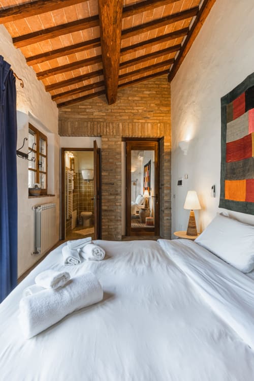 Camera rustica con travi in legno e dettagli in mattone dell'hotel Buonconvento, letti gemelli con biancheria bianca e asciugamani, bagno privato