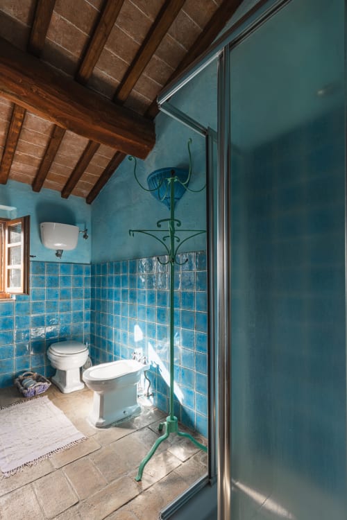 Bagno rustico con piastrelle turchesi, sanitari bianchi, travi in legno a vista e appendiabiti verde nella struttura di Buonconvento
