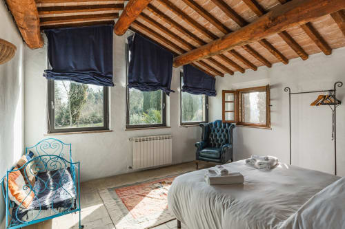 Camera da letto rustica a Buonconvento con travi in legno a vista, letto in ferro blu, poltrona in pelle e vista sul giardino