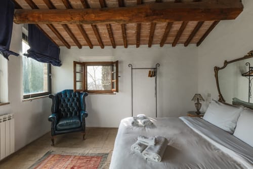Camera rustica con travi in legno a vista, poltrona in pelle blu, letto bianco e finestra aperta a Buonconvento, Italia