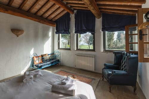 Camera da letto in rustico toscano con travi in legno, camino, poltrone blu scuro e vista sulla campagna a Buonconvento