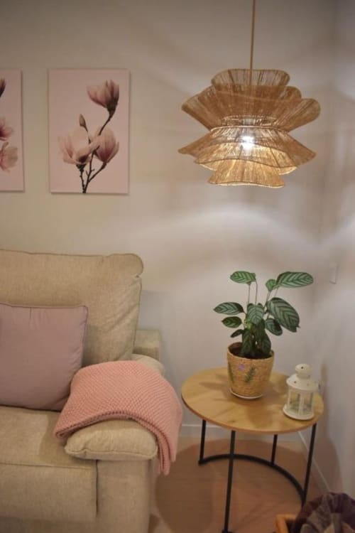 Rincón acogedor con sofá beige, manta rosa, maceta sobre mesa de madera, cuadro floral y lámpara colgante tejida