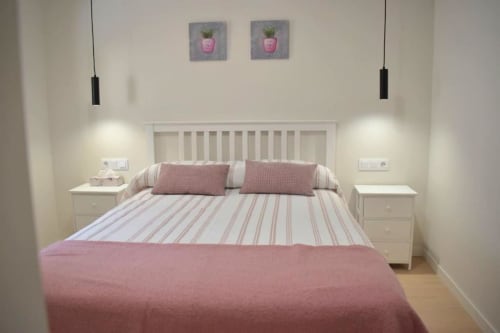 Dormitorio luminoso con cama de rayas blancas, almohadas rosas, lámparas colgantes y dos cuadros de flores en maceta en Casa El Abuelo rural