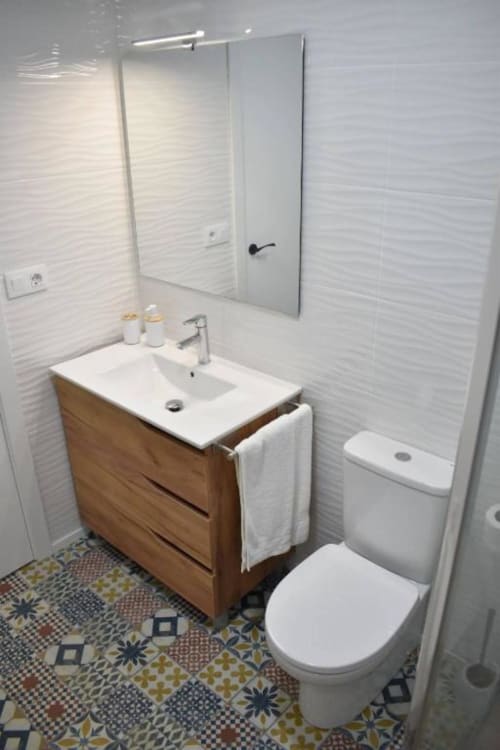 Baño moderno con azulejos blancos texturizados, mueble de madera, lavabo blanco, inodoro, espejo y azulejos decorados en el piso de Casa Rural