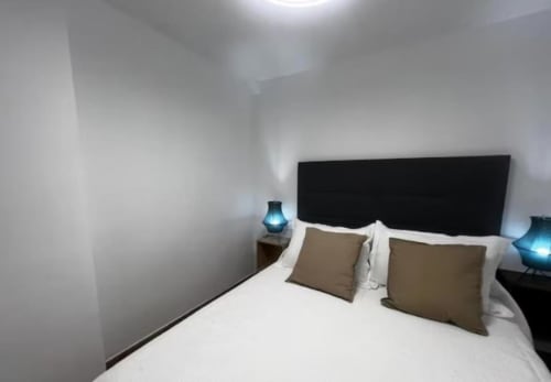 Dormitorio moderno en apartamento Las Arenas de València con cabecero negro, ropa de cama blanca, almohadas beige y lámparas azules