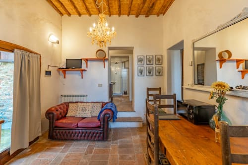Accogliente soggiorno di appartamento toscano con divano Chesterfield, travi in legno, pavimenti in cotto e vista sul giardino a Buonconvento