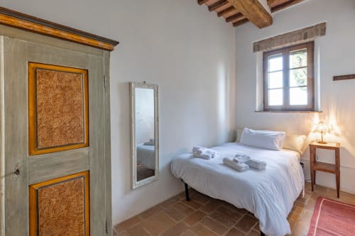 Camera da letto rustica italiana con biancheria bianca, travi in legno, porta in sughero, specchio, finestra, lampada comodino e pavimenti in cotto