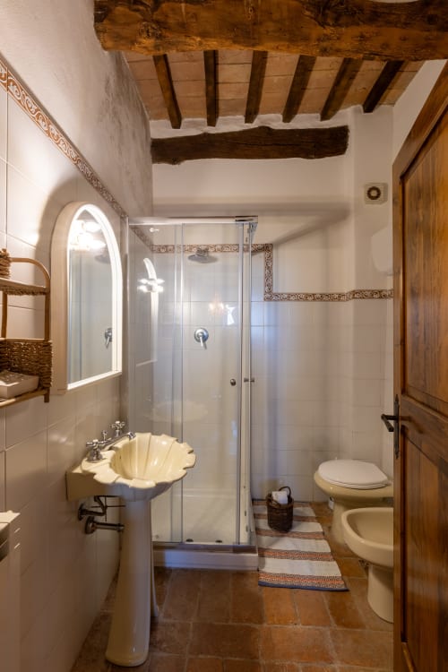 Bagno rustico dell'hotel Quarantallina a Buonconvento con lavabo in conchiglia, box doccia in vetro e travi in legno