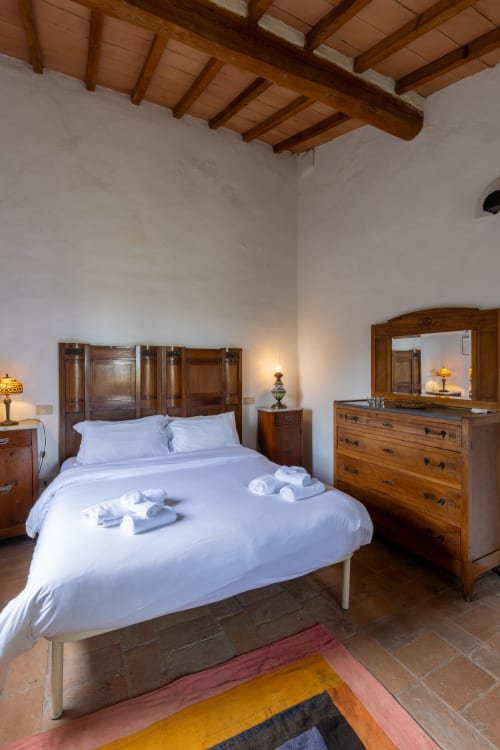 Camera rustica dell'hotel Buonconvento con soffitto in travi di legno, biancheria bianca, mobili in legno e tappeto colorato