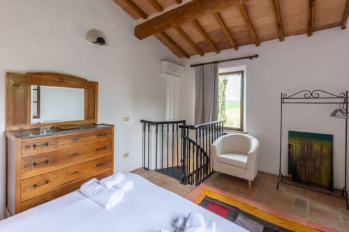Camera da letto rustica con travi in legno, pavimenti in terracotta, letto bianco, cassettone in legno e poltrona color crema a Buonconvento, Toscana.