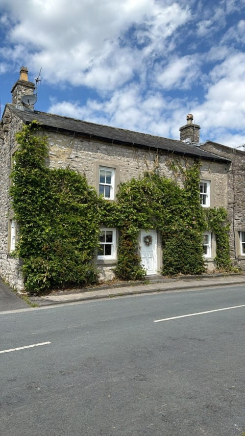 Ivy-covered stone cottage in Austwick with white door, windows, and chimneys under blue sky