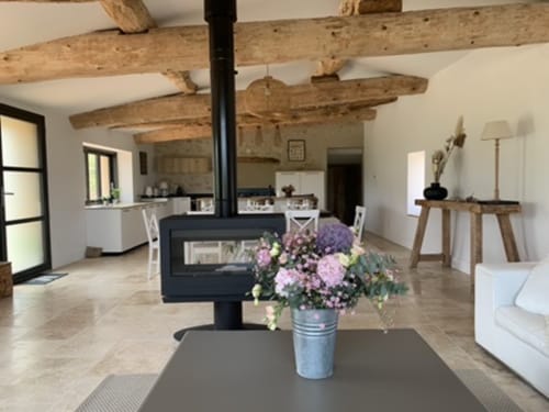Intérieur de chaumière ouverte avec poutres en bois apparentes, poêle à bois, fleurs roses en vase, mobilier blanc et cuisine en Drôme