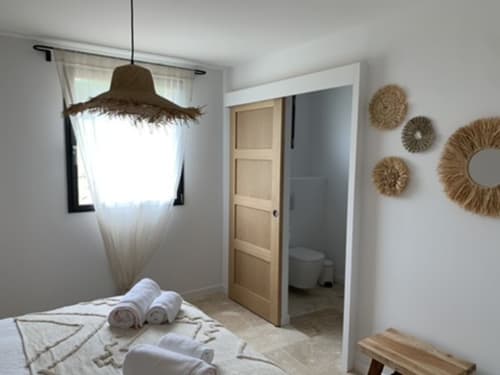 Chambre lumineuse dans un gîte de la Drôme avec luminaire suspendu en osier, draps blancs, serviettes roulées et décoration murale en paille.