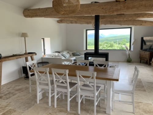 Salle à manger ouverte de cottage moderne avec vue sur les montagnes, poutres en bois, cheminée et chaises blanches à table en bois dans la Drôme
