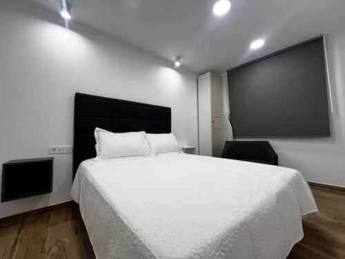 Moderno dormitorio en el apartamento Las Arenas de València con cabecero negro, ropa de cama blanca, suelo de madera y decoración minimalista