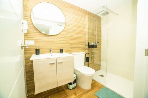 Baño moderno del apartamento Las Arenas con pared de madera, espejo redondo, lavamanos y ducha abierta