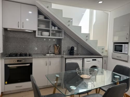 Modern kitchen-dining area in Chaves apartment with white cabinets, gray counters, glass table, and integrated laundry