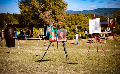 Exposition d'art en plein air avec des portraits sur chevalets dans un champ herbeux, montagnes et arbres en arrière-plan