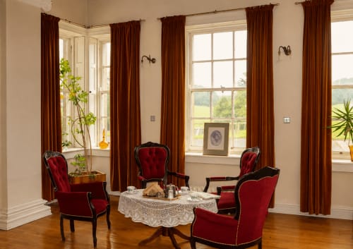 Salon élégant de South Hill House avec fauteuils en velours rouge, table ronde et rideaux bordeaux donnant sur la campagne