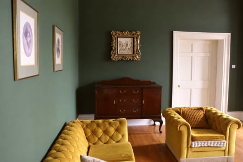 Élégant salon vert de South Hill House avec fauteuils jaunes capitonnés, buffet en bois foncé et tableaux encadrés