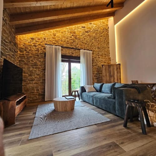 Suite Izarbe con pared de piedra, vigas de madera, sofá azul y puertas al balcón con vista a la naturaleza
