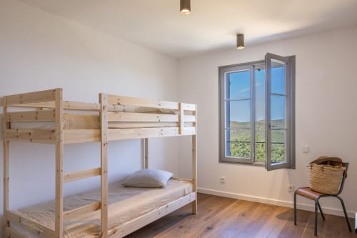 Chambre lumineuse avec lits superposés en bois, grande fenêtre vue sur les collines d'Ersa, plancher en bois et fauteuil en rotin dans villa