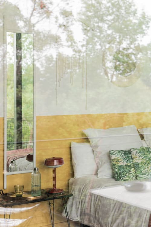 Chambre lumineuse à la Maison Jalon avec tête de lit dorée, literie blanche, coussin plante verte et vue fenêtre encadrée d'arbres
