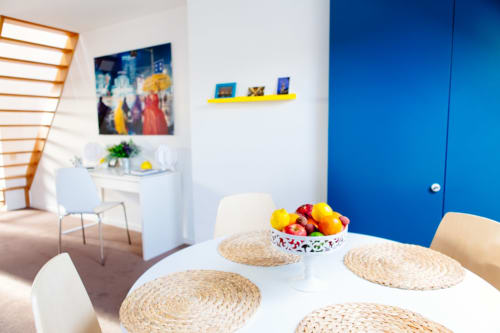 Salle à manger moderne dans un studio B&B bruxellois avec table blanche, sets de table tressés, coupe de fruits frais et porte bleu vif